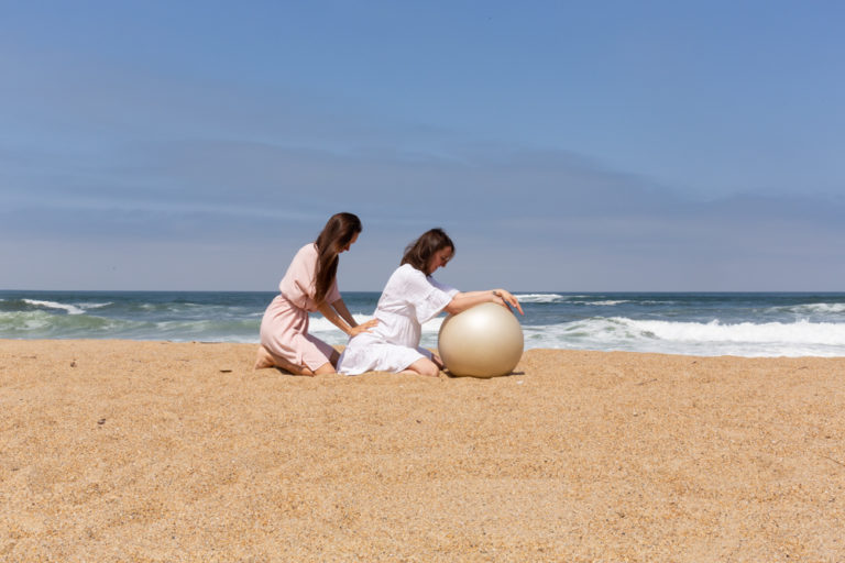 Monika working with a pregnant client as a doula on Biarritz beach, the client is leaning on the pilates ball